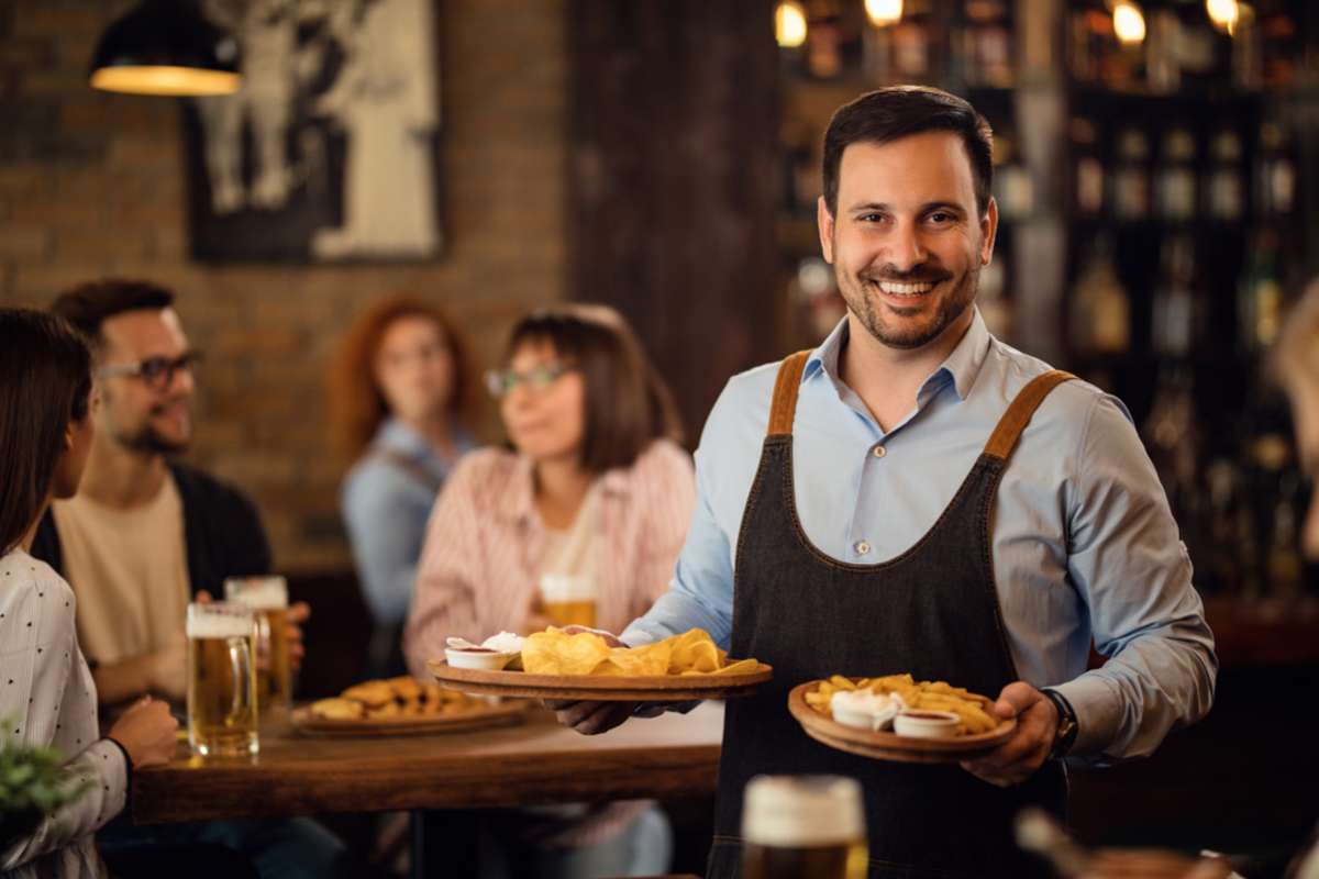 Happy waiter holding plates with food and looking at camera while serving guests in a restaurant
