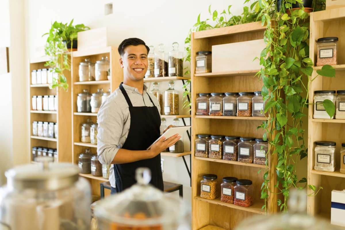 Portrait of a happy young man and business owner smiling while making eye contact and standing inside the bulk shop