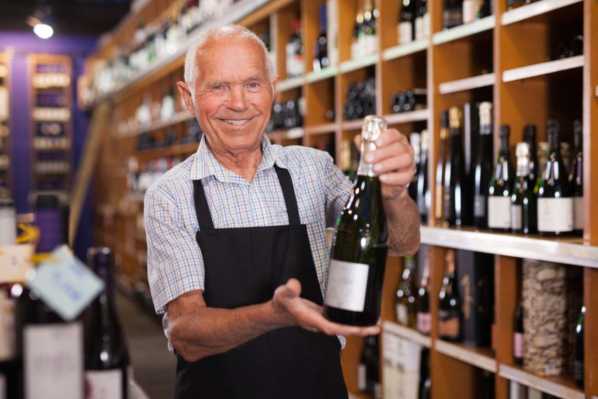 Portrait of confident elderly male owner of wine shop offering to buy wine bottle