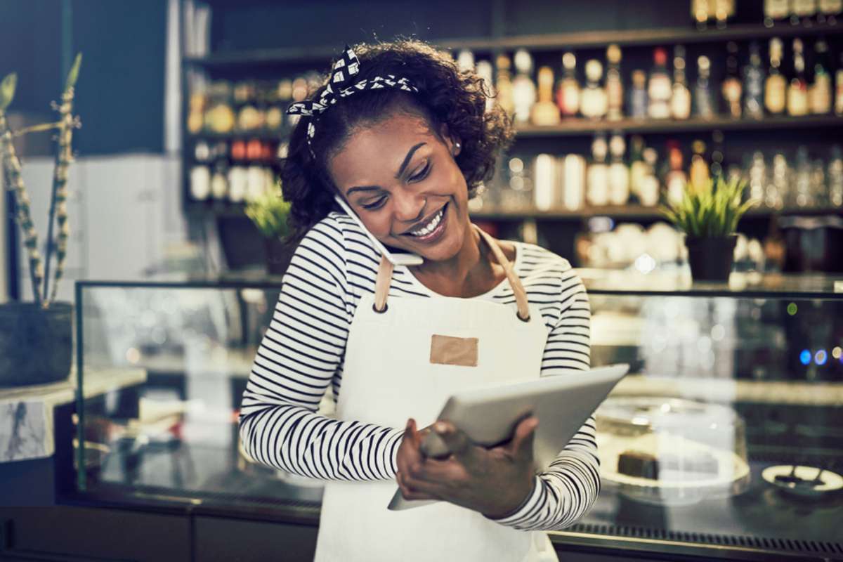 Smiling young African entrepreneur standing in front of the counter of her cafe talking on a cellphone and using a tablet