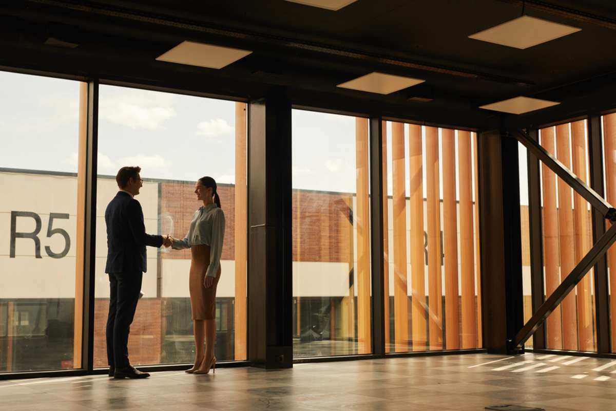 Wide angle view of real estate agent shaking hands with client while standing in empty office building interior lit by sunlight