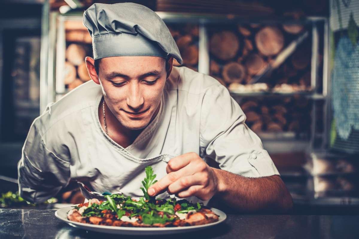Young smiling chef in white uniform and gray hat decorate ready dish with green rucola herbs in interior of modern restaurant kitchen
